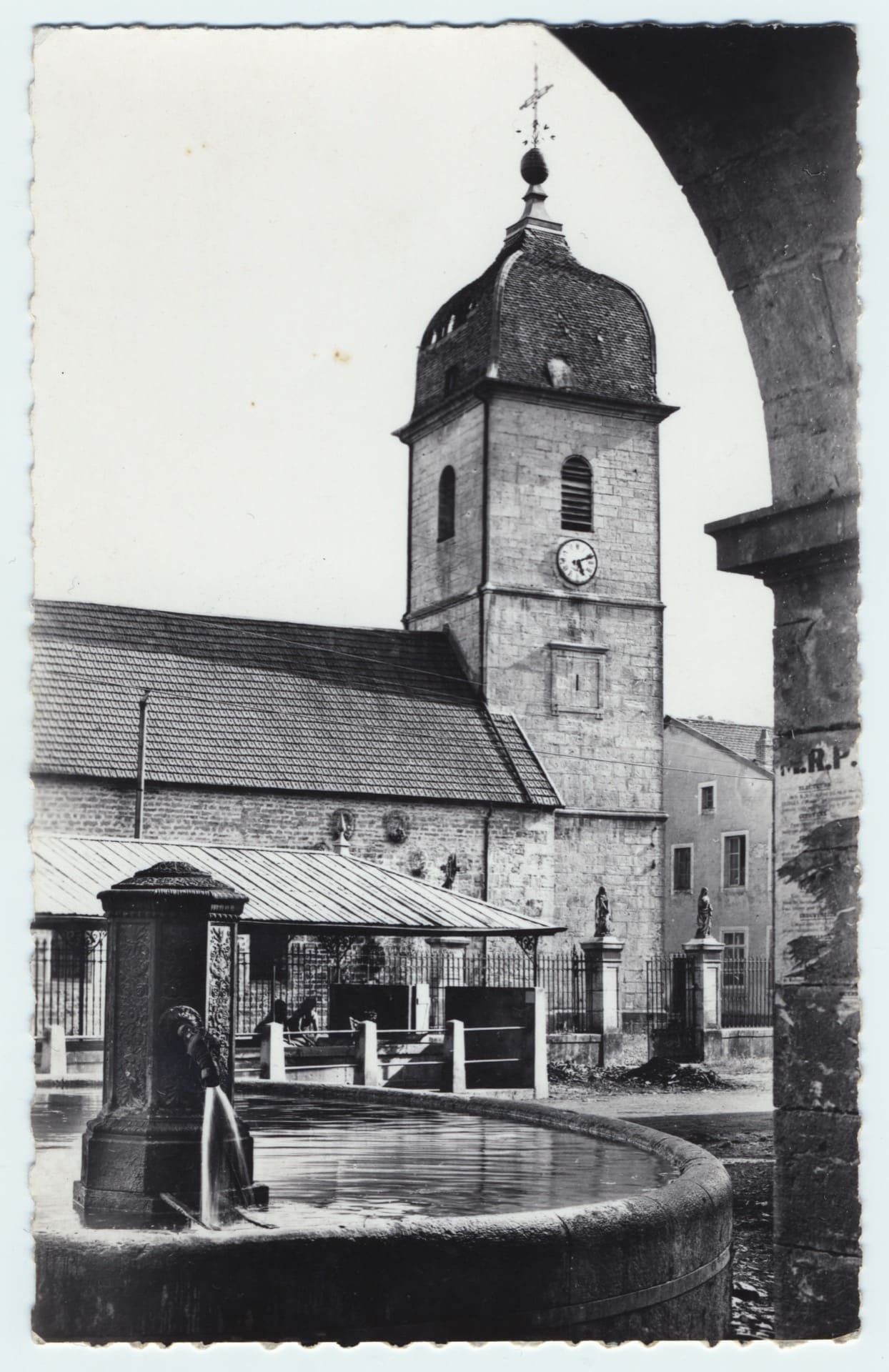 Carte postale ancienne verticale de la Place de l'Église à Landresse. L'image est cadrée depuis l'arche en pierre de la mairie, montrant une fontaine au premier plan et le clocher comtois en arrière-plan.