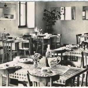 Photographie ancienne de la salle à manger de l'Hôtel-Restaurant Cretin à Landresse. Les tables sont prêtes avec des nappes à carreaux, de la vaisselle, un cendrier Cinzano et un pichet publicitaire de la distillerie Émile Pernot de Pontarlier.