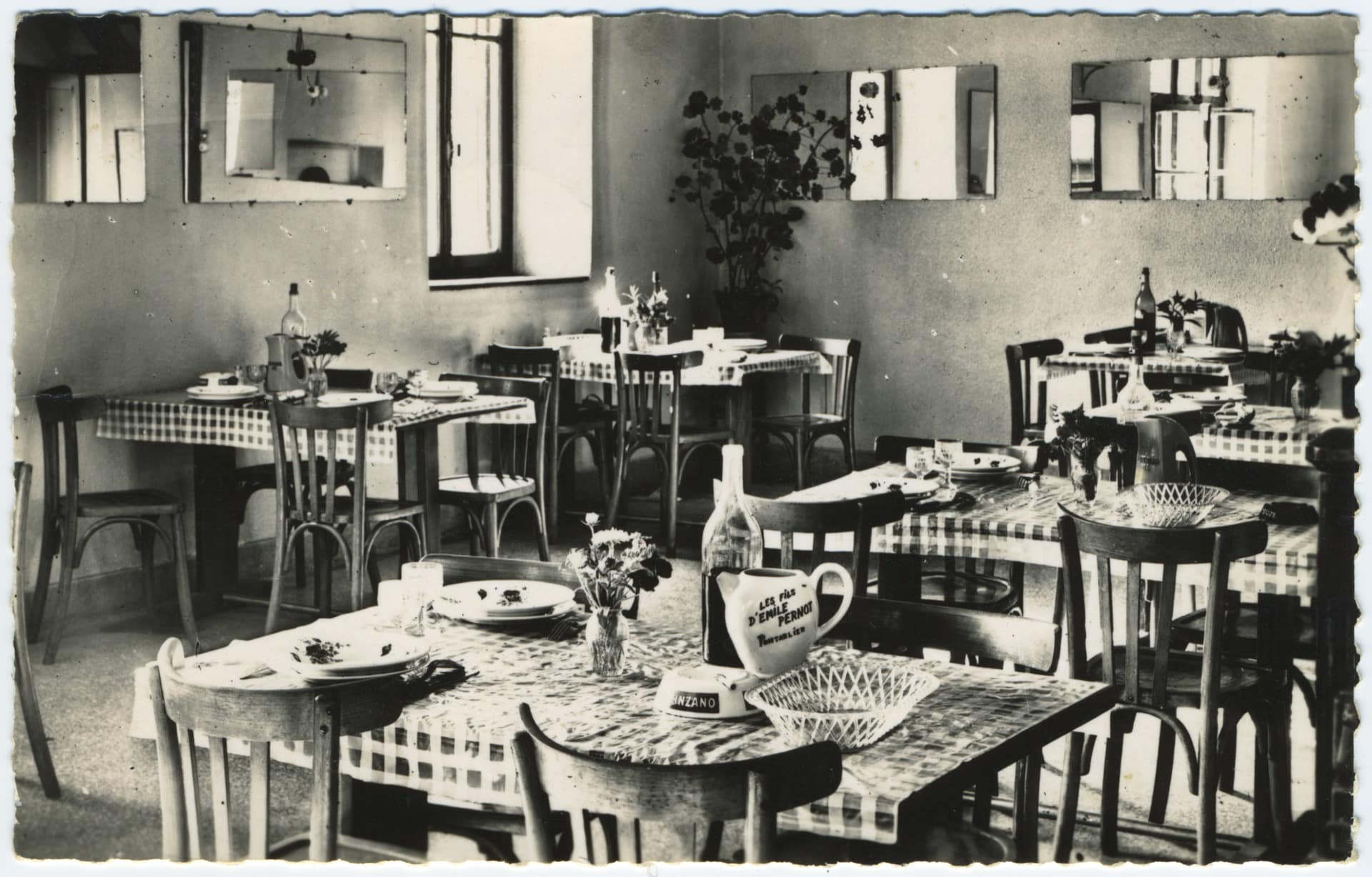 Photographie ancienne de la salle à manger de l'Hôtel-Restaurant Cretin à Landresse. Les tables sont prêtes avec des nappes à carreaux, de la vaisselle, un cendrier Cinzano et un pichet publicitaire de la distillerie Émile Pernot de Pontarlier.