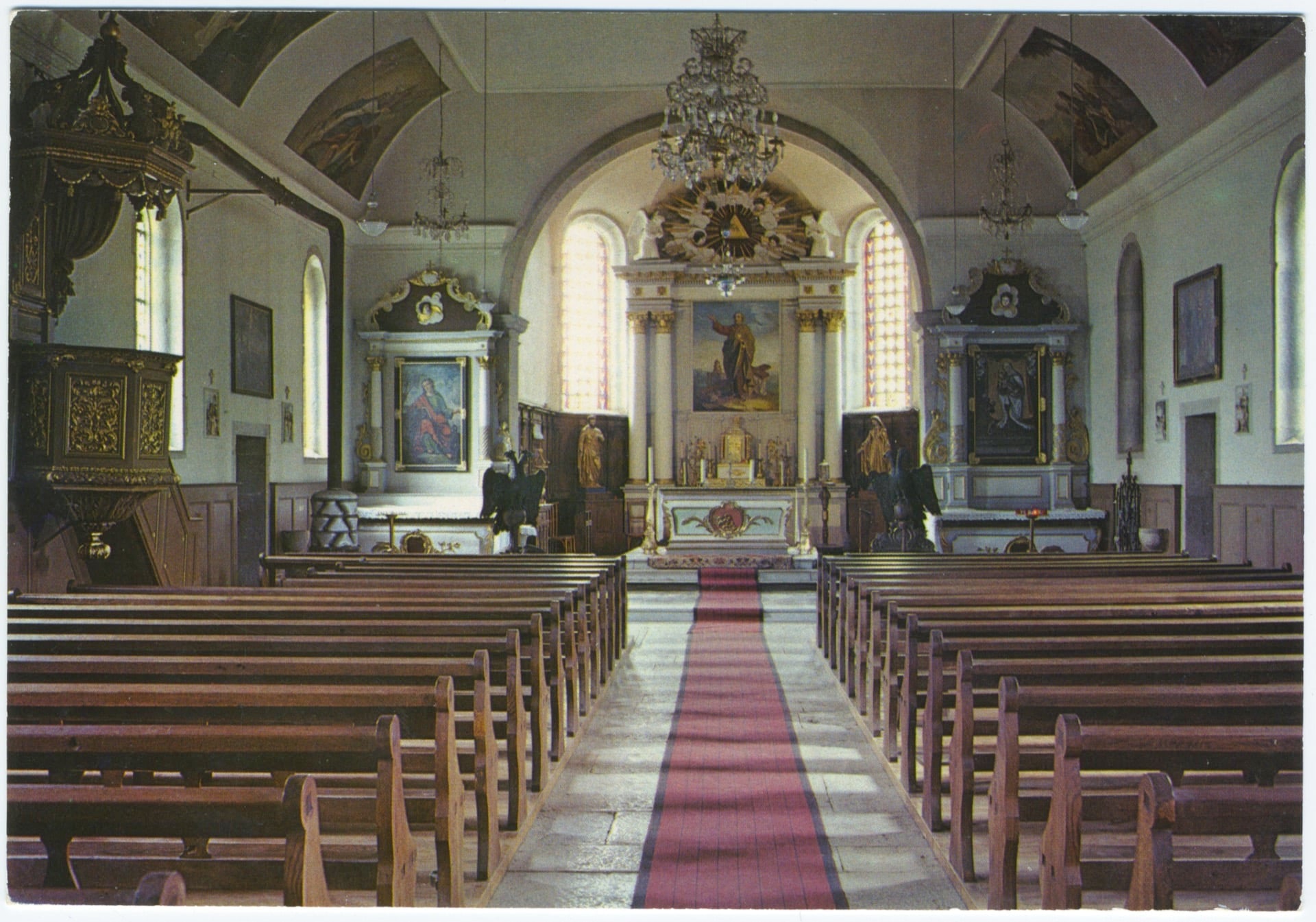 Photographie en couleur de l'intérieur de l'église de Landresse. L'allée centrale avec un tapis rouge sépare les bancs en bois et mène vers le chœur richement décoré d'autels baroques blancs et dorés sous une voûte peinte.